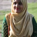 A young woman in traditional hijab attire smiles warmly, showcasing cultural fashion outdoors.